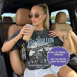 Pu&ograve; includere: T-shirt grigio scuro con grafica "THE HOLLOW", uno striscione con la scritta "Inn for Travelers and Adventurers" e "MERRYWOOD FOREST ESTD 2021". La maglietta &egrave; indossata con una gonna bianca e gioielli d'oro. Viene tenuta una bevanda al caff&egrave;.