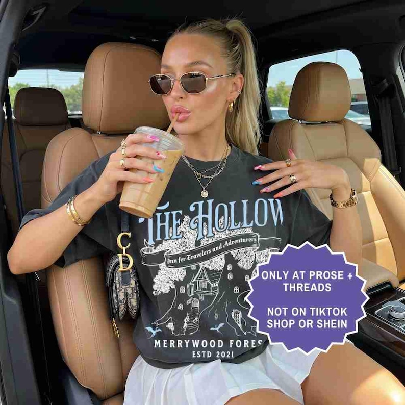 Pu&ograve; includere: T-shirt grigio scuro con grafica "THE HOLLOW", uno striscione con la scritta "Inn for Travelers and Adventurers" e "MERRYWOOD FOREST ESTD 2021". La maglietta &egrave; indossata con una gonna bianca e gioielli d'oro. Viene tenuta una bevanda al caff&egrave;.