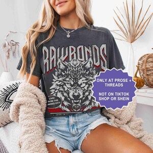 Op de afbeelding: Donkergrijs t-shirt met een wolfafbeelding en de tekst "RAWRONDS". Het shirt heeft ook de tekst "LIVE! ROM THE INDED CITY". Een paars bord zegt "ONLY AT PROSE + THREADS NOT ON TIKTOK SHOP OR SHEIN".