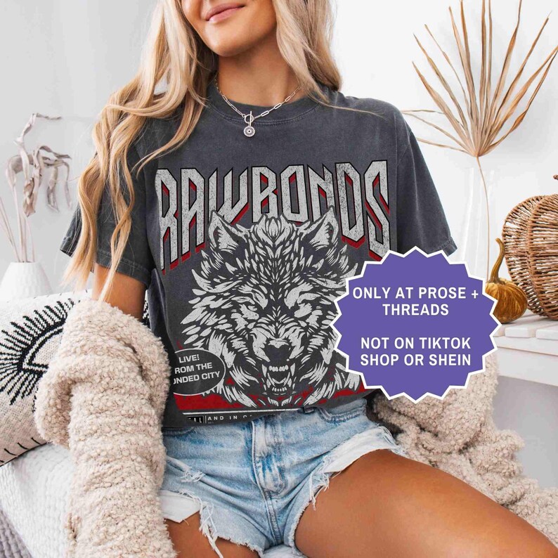 Op de afbeelding: Donkergrijs t-shirt met een wolfafbeelding en de tekst "RAWRONDS". Het shirt heeft ook de tekst "LIVE! ROM THE INDED CITY". Een paars bord zegt "ONLY AT PROSE + THREADS NOT ON TIKTOK SHOP OR SHEIN".