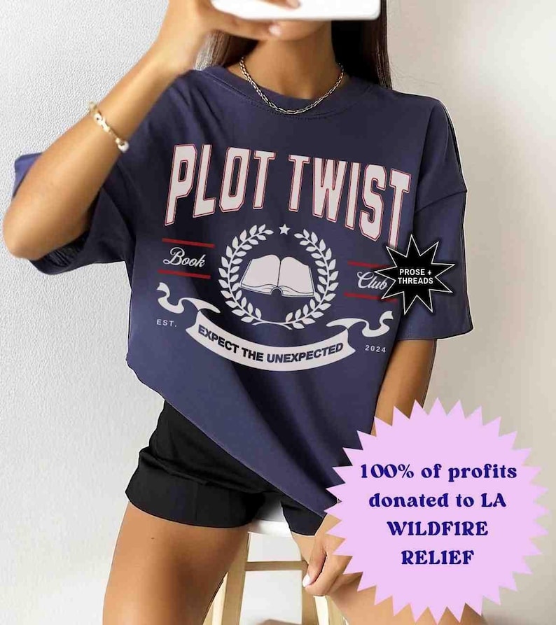 May include: A navy blue t-shirt with the text "PLOT TWIST" in white with red outlines. The shirt features a white graphic of a book with a laurel wreath and the text "Book Club" and "EST. 2024". The text "PROSE + THREADS" is in a white starburst. The shirt is being worn by a person with black shorts. The text "100% of profits donated to LA WILDFIRE RELIEF" is in a purple starburst.