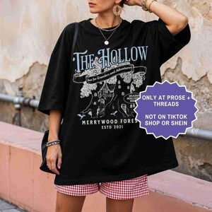 Pu&ograve; includere: T-shirt nera oversize con un design grafico con il testo "The Hollow" e "Merrywood Fores ESTD 2021". Il design include un albero con una casa e le parole "Inn for Travelers and Adventurers". Un adesivo viola dice "ONLY AT PROSE + THREADS NOT ON TIKTOK SHOP OR SHEIN".