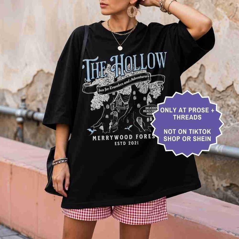 Pu&ograve; includere: T-shirt nera oversize con un design grafico con il testo "The Hollow" e "Merrywood Fores ESTD 2021". Il design include un albero con una casa e le parole "Inn for Travelers and Adventurers". Un adesivo viola dice "ONLY AT PROSE + THREADS NOT ON TIKTOK SHOP OR SHEIN".