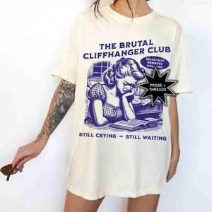 May include: Cream-colored t-shirt with a purple graphic. The graphic features a vintage illustration of a woman reading and crying, with text that reads "THE BRUTAL CLIFFHANGER CLUB" and "STILL CRYING - STILL WAITING."