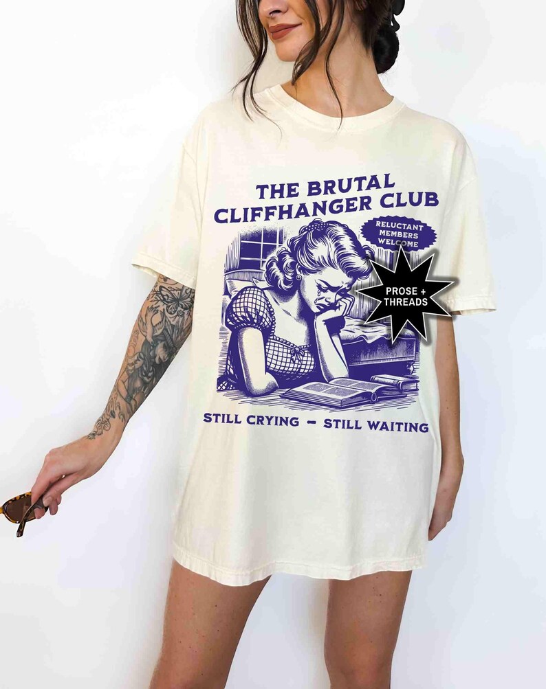May include: Cream-colored t-shirt with a purple graphic. The graphic features a vintage illustration of a woman reading and crying, with text that reads "THE BRUTAL CLIFFHANGER CLUB" and "STILL CRYING - STILL WAITING."