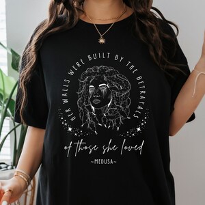 May include: Black t-shirt featuring a white illustration of Medusa with snakes for hair, surrounded by text that reads "HER WALLS WERE BUILT BY THE BETRAYALS of those she loved - MEDUSA-". The design is centered on the shirt.