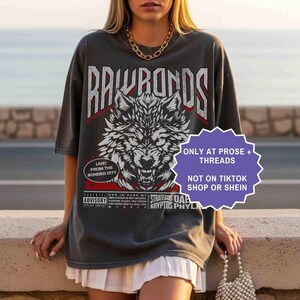 Op de afbeelding: Donkergrijs t-shirt met een wolfafbeelding en de tekst "RAWRONDS". Het shirt heeft ook de tekst "LIVE! FROM THE BONDED CITY" en "STRATEGOS DAA KRYPTOS PHYL". Een paars bord zegt "ONLY AT PROSE + THREADS NOT ON TIKTOK SHOP OR SHEIN".
