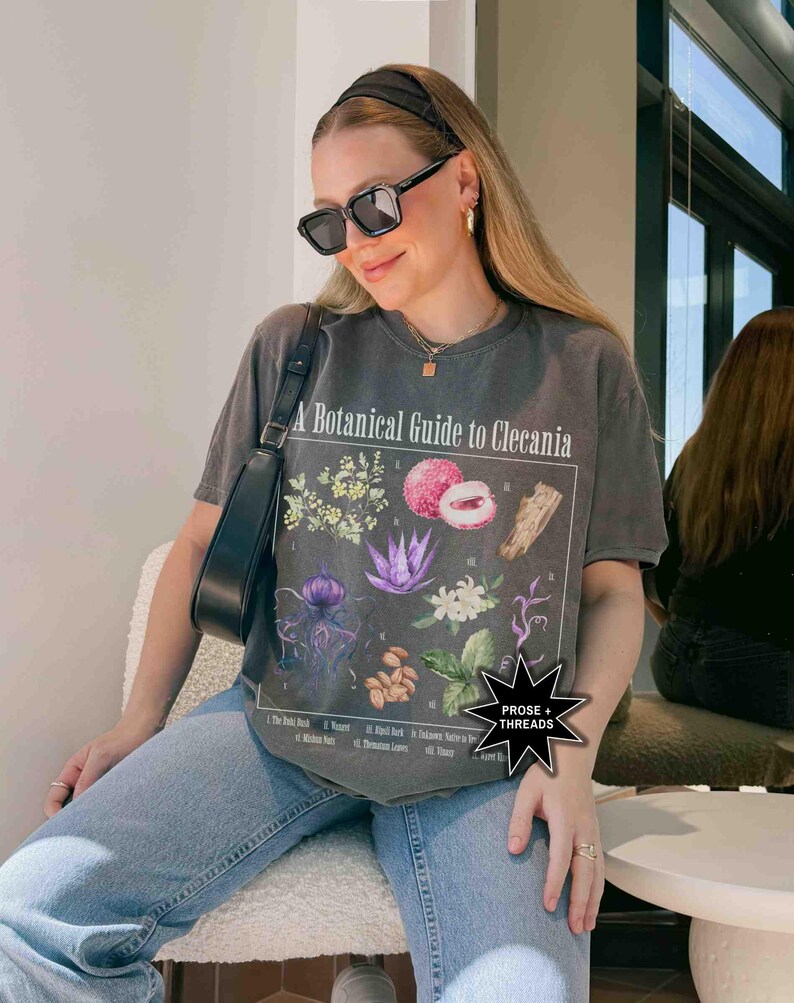 May include: A charcoal grey t-shirt with a botanical illustration and the text "A Botanical Guide to Clecania". The illustration features various plants and flowers, including a lychee fruit, a flower with white petals, and a purple flower. The t-shirt has a relaxed fit and short sleeves.