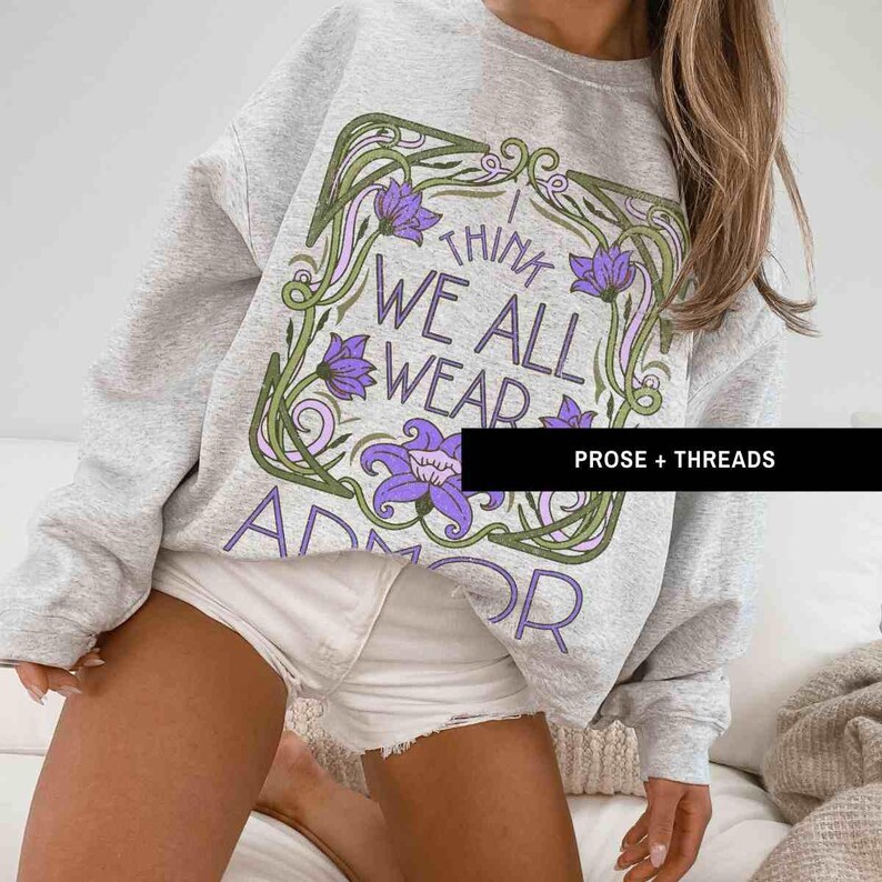 May include: A gray sweatshirt with a floral design and the text "I think we all wear armor" printed on the front. The sweatshirt is being worn by a person with long brown hair.