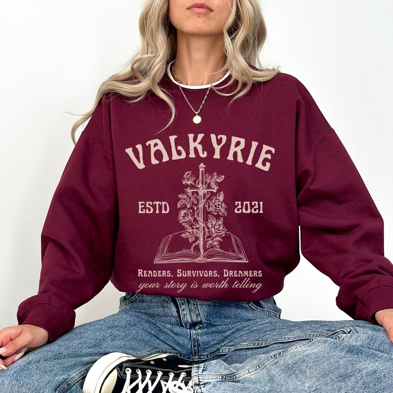 Może przedstawiać: Bordowa bluza z białym nadrukiem graficznym, na kt&oacute;rym widnieje napis "Valkyrie Est. 2021" z mieczem i r&oacute;żami. Tekst pod grafiką brzmi "Readers, Survivors, Dreamers your story is worth telling."
