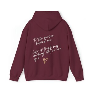 May include: Burgundy hoodie with a hood and long sleeves. The back of the hoodie features the text "To the person behind me, Life is tough my darling, but so are you" with a gold heart symbol.