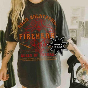 Pode incluir: T-shirt cinzento escuro com o texto "BEGIN GALATHYNID FIREHEAR QUEEN OF TERAS" em laranja e vermelho. A camisola tem um aspeto vintage e desgastado. O texto inclui as frases "ATTLE THE STARS" e "ADARLAN'S ASSASSIN".