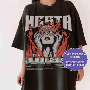 May include: Black t-shirt with a graphic design featuring a woman with a mask, surrounded by flames, and the word "MERTA" above. The shirt also has the text "CAULDRON BLESSED" and other text below the graphic.