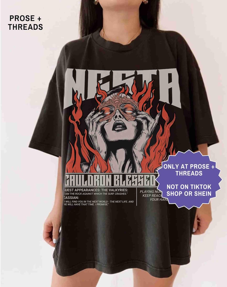 May include: Black t-shirt with a graphic design featuring a woman with a mask, surrounded by flames, and the word "MERTA" above. The shirt also has the text "CAULDRON BLESSED" and other text below the graphic.
