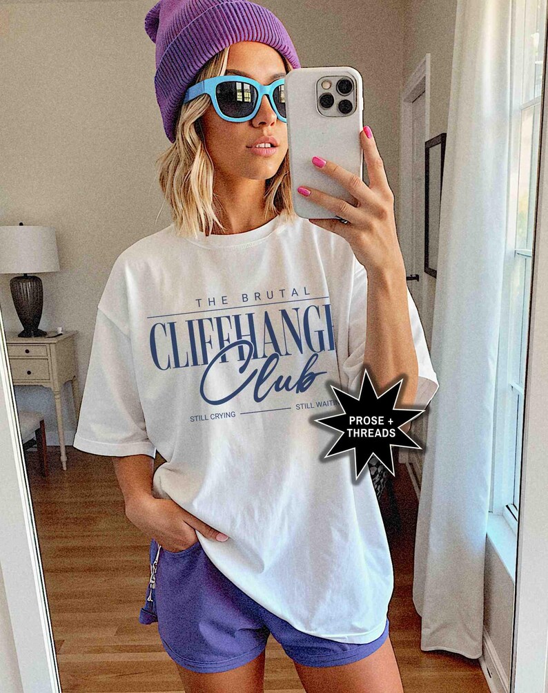 May include: A white t-shirt with the text "The Brutal Cliffhanger Club" in blue lettering. The text "Still Crying" and "Still Waiting" are printed below the main text. The t-shirt is being worn by a person with a purple beanie and blue sunglasses. The image also includes a black star with the text "Prose + Threads" in white lettering.