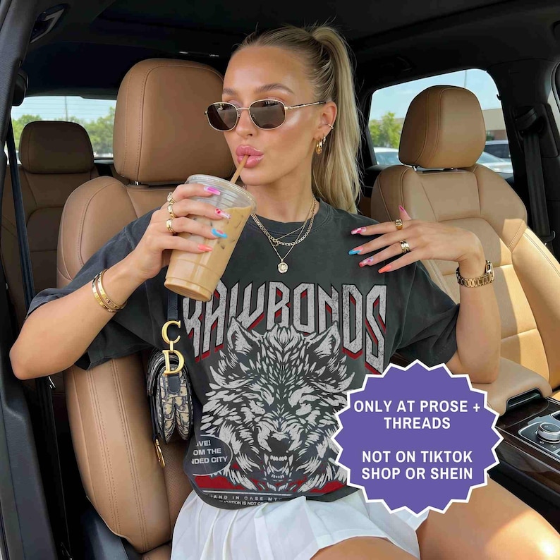 Op de afbeelding: Een vrouw draagt een donkergrijs grafisch t-shirt met een wolfontwerp en de tekst "RAWBONDS". Ze draagt een zonnebril en sieraden en houdt een drankje vast. De afbeelding bevat een witte rok en een kleine handtas. Een paars bord zegt "ONLY AT PROSE + THREADS NOT ON TIKTOK SHOP OR SHEIN".