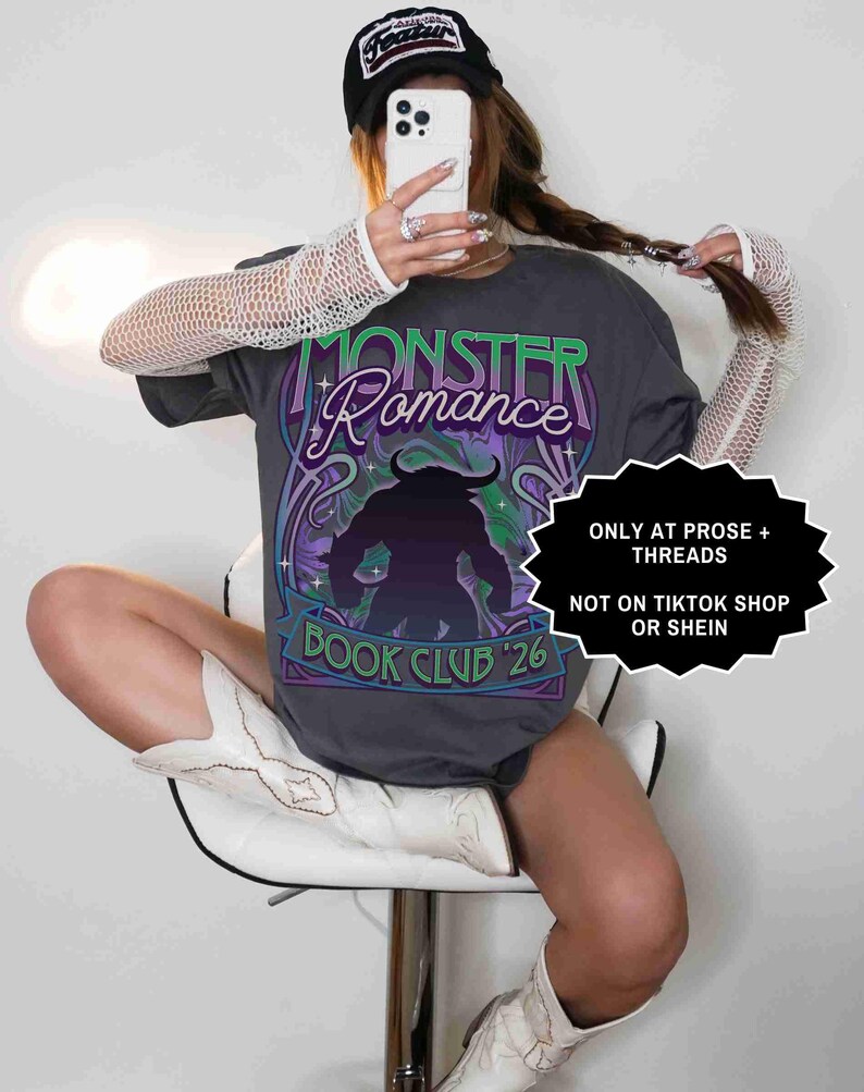 Op de afbeelding: Donkergrijs t-shirt met de tekst "MONSTER Romance BOOK CLUB '26" in groen en paars. Het shirt heeft een monsterlijke afbeelding. De afbeelding bevat ook een zwarte pet en witte laarzen.