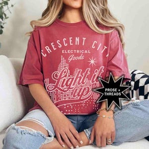 May include: A red t-shirt with a vintage-style graphic design that says "Crescent City Electrical Goods Light it Up" with a cityscape illustration. The graphic is in white and black. The t-shirt is worn by a person sitting on a couch.