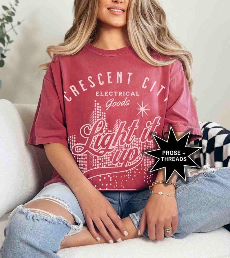 May include: A red t-shirt with a vintage-style graphic design that says "Crescent City Electrical Goods Light it Up" with a cityscape illustration. The graphic is in white and black. The t-shirt is worn by a person sitting on a couch.