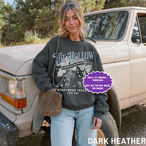 May include: Dark heather grey sweatshirt featuring "THE HOLLOW" graphic, a tree illustration, and "Merrywood Forest Estd 2021" text. The sweatshirt is paired with light blue jeans and a patterned purse. A vintage truck is in the background.