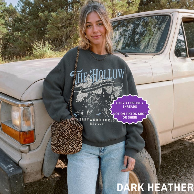 May include: Dark heather grey sweatshirt featuring "THE HOLLOW" graphic, a tree illustration, and "Merrywood Forest Estd 2021" text. The sweatshirt is paired with light blue jeans and a patterned purse. A vintage truck is in the background.