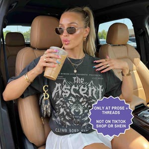 May include: A woman wearing sunglasses and a black graphic t-shirt with the text "The Ascent" and a wolf design. She is holding a beverage in a clear plastic cup with a straw. The shirt also has the text "CLIMB BOND" and "AUG 2025".