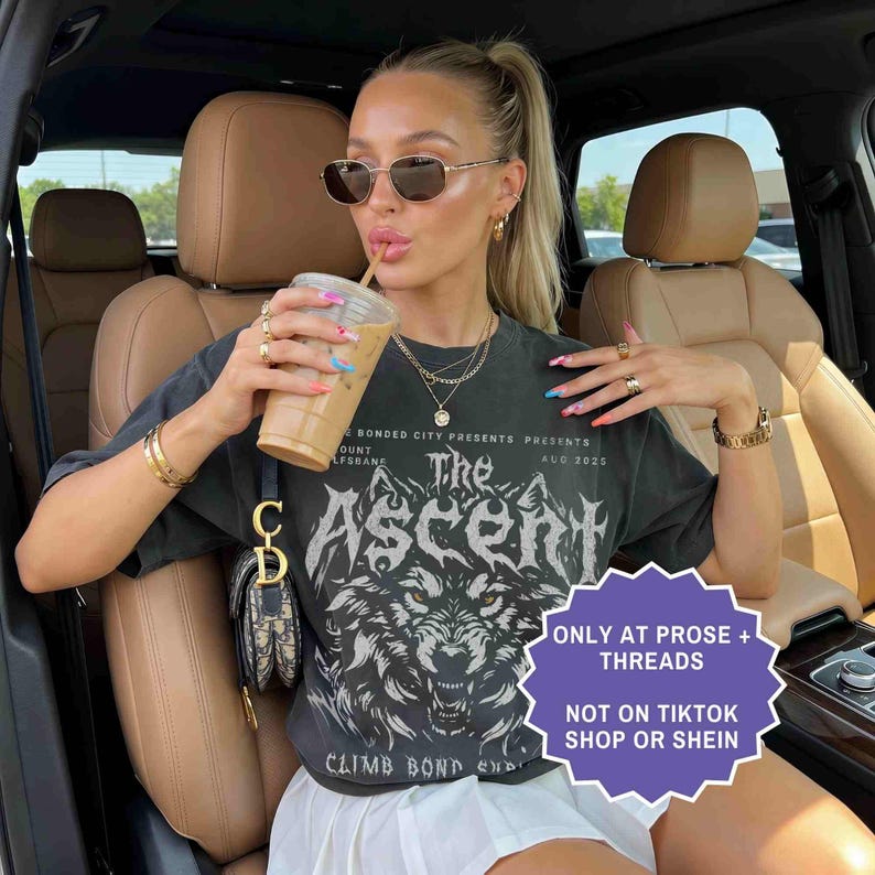 May include: A woman wearing sunglasses and a black graphic t-shirt with the text "The Ascent" and a wolf design. She is holding a beverage in a clear plastic cup with a straw. The shirt also has the text "CLIMB BOND" and "AUG 2025".