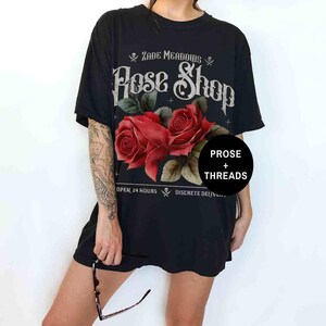May include: Black t-shirt with a graphic of a rose shop called "Zade Meadows Rose Shop". The graphic features two red roses with green leaves and a skull and crossbones on either side of the shop name. The text "Prose + Threads" is in a circle below the roses.