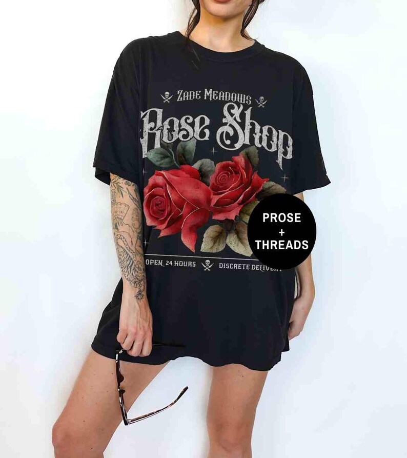 May include: Black t-shirt with a graphic of a rose shop called "Zade Meadows Rose Shop". The graphic features two red roses with green leaves and a skull and crossbones on either side of the shop name. The text "Prose + Threads" is in a circle below the roses.
