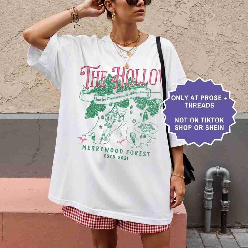 Pu&ograve; includere: T-shirt bianca con una grafica che raffigura la locanda "The Hollow", un design di casa sull'albero e la scritta "Merrywood Forest ESTD 2021". La maglietta ha anche un'etichetta viola con la scritta "ONLY AT PROSE + THREADS NOT ON TIKTOK SHOP OR SHEIN."