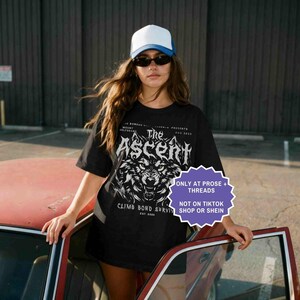 May include: Black t-shirt with a wolf graphic and the text "The Ascent". The shirt also includes the text "Climb Bond Survive" and "Est 2025". The person in the image is wearing a white and blue trucker hat and sunglasses.