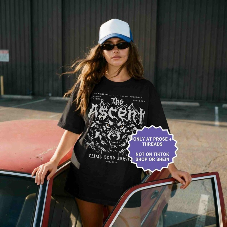 May include: Black t-shirt with a wolf graphic and the text "The Ascent". The shirt also includes the text "Climb Bond Survive" and "Est 2025". The person in the image is wearing a white and blue trucker hat and sunglasses.