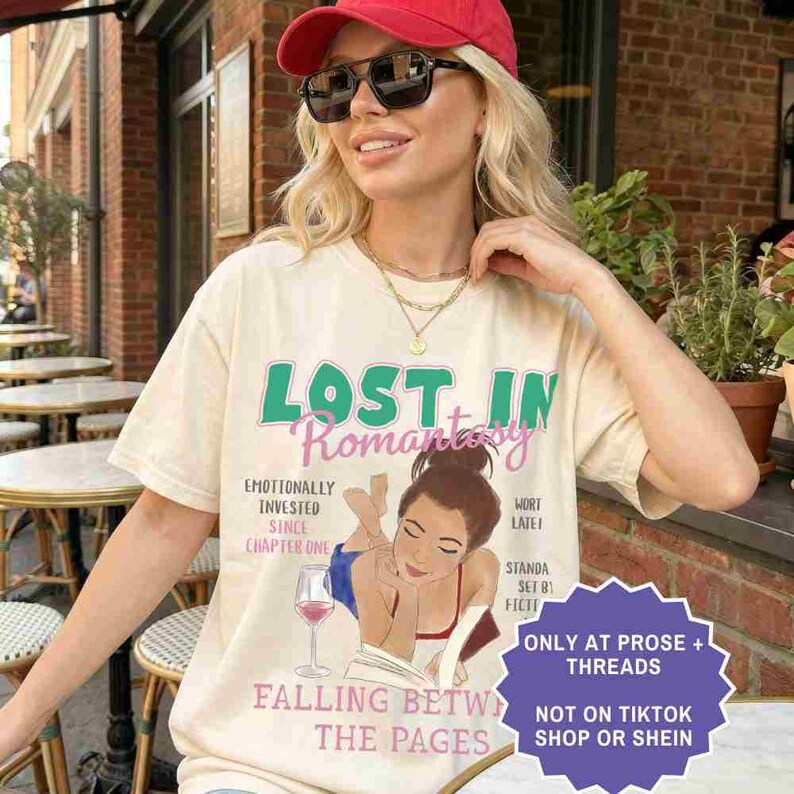 May include: Cream-colored t-shirt with the text "LOST IN Romantasy" in green and pink. The shirt features an illustration of a woman reading a book with a glass of wine. Additional text includes "Emotionally Invested Since Chapter One" and "Falling Between The Pages."