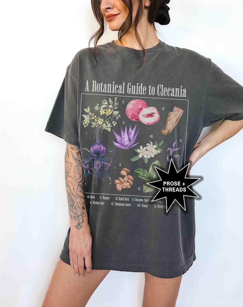 May include: A dark gray t-shirt with a vintage-style graphic print of a botanical illustration. The graphic features a variety of plants and fruits, including a lychee, a flower, and a leaf. The text "A Botanical Guide to Clecania" is printed above the illustration. The text "PROSE + THREADS" is printed in a star shape on the bottom right corner of the shirt.