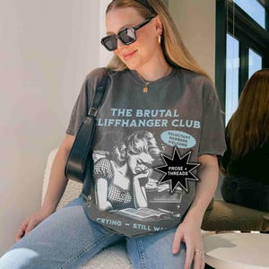 May include: Dark gray t-shirt with a distressed graphic of a crying woman and the text "THE BRUTAL LIFFHANGER CLUB." The shirt also features the text "Crying - Still Writing" and "Reluctant Members Welcome."
