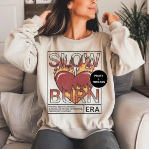 May include: A beige sweatshirt with a graphic print of a heart with flames and the text "SLOW BURN" in a vintage style. The text "PROSE + THREADS" and "ERA" are also included in the design.