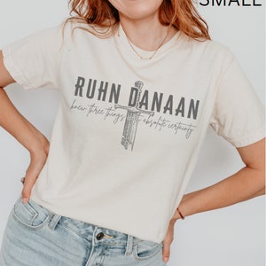 May include: A cream coloured t-shirt with a graphic of a sword and the text "Ruhn Danaan knew three things with absolute certainty".