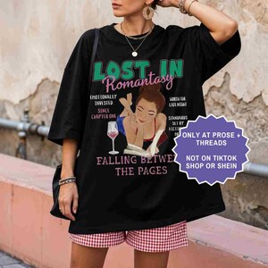 May include: Black oversized t-shirt with the text "Lost in Romantasy" in green and pink. The shirt features an illustration of a woman reading with a glass of wine. Additional text includes "Falling Between the Pages."