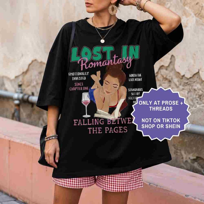 May include: Black oversized t-shirt with the text "Lost in Romantasy" in green and pink. The shirt features an illustration of a woman reading with a glass of wine. Additional text includes "Falling Between the Pages."