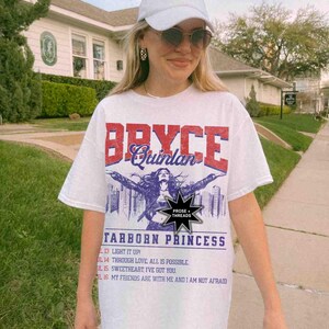 May include: White t-shirt featuring a graphic print of a woman with the text "Bryce Quinlan" and "Starborn Princess". The graphic also includes the text "Prose + Threads".