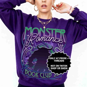 May include: A purple sweatshirt with the text "MONSTER Romance BOOK CLUB" in green and white. A black silhouette of a monster is in the center. The sweatshirt has a crew neck and long sleeves.