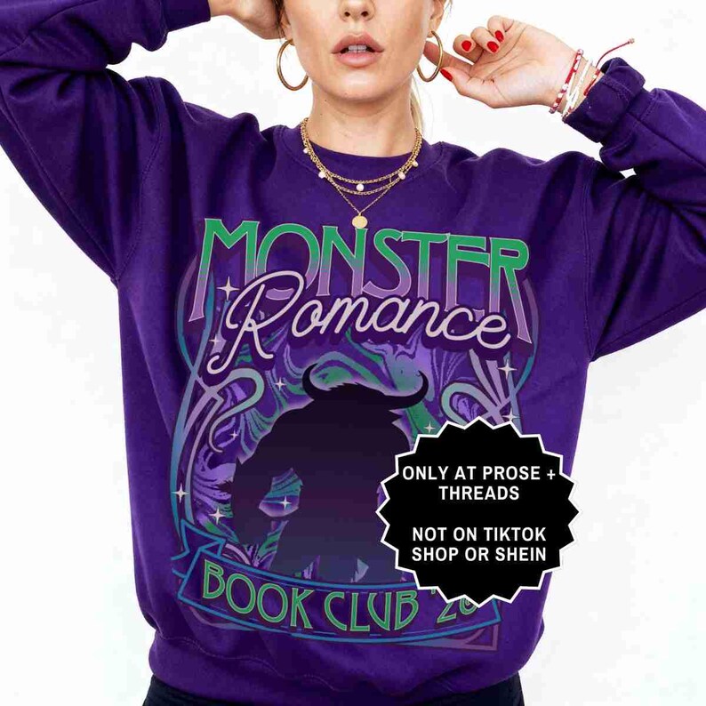 May include: A purple sweatshirt with the text "MONSTER Romance BOOK CLUB" in green and white. A black silhouette of a monster is in the center. The sweatshirt has a crew neck and long sleeves.