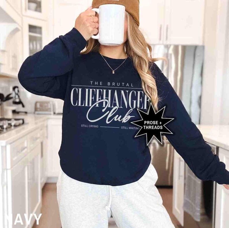 Puede incluir: Sudadera de manga larga azul marino con el texto "The Brutal Cliffhanger Club" en blanco. Los textos "Still Crying" y "Still Waiting" est&aacute;n impresos debajo del texto principal. Una estrella negra con el texto "Prose + Threads" est&aacute; impresa en el lado derecho de la sudadera.