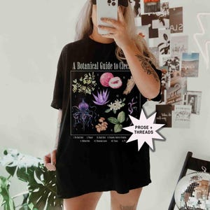 May include: Black t-shirt with a botanical illustration and the text "A Botanical Guide to Clec." The illustration features various plants and flowers, including a lychee, a jellyfish, and a flower with purple petals. The text "PROSE + THREADS" is in a white starburst.