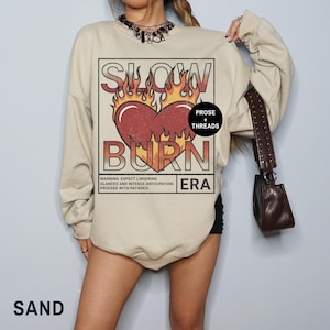 May include: A beige sweatshirt with a graphic print of a heart with flames and the text "Slow Burn Era" in a vintage style. The text "Prose + Threads" is also included in the graphic.
