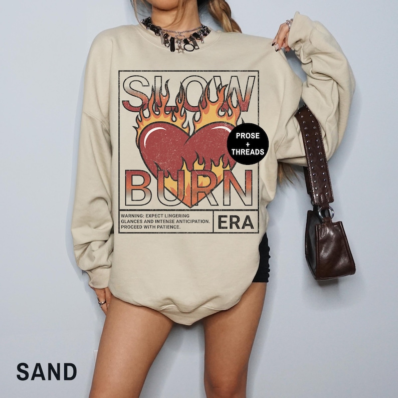 May include: A beige sweatshirt with a graphic print of a heart with flames and the text "Slow Burn Era" in a vintage style. The text "Prose + Threads" is also included in the graphic.