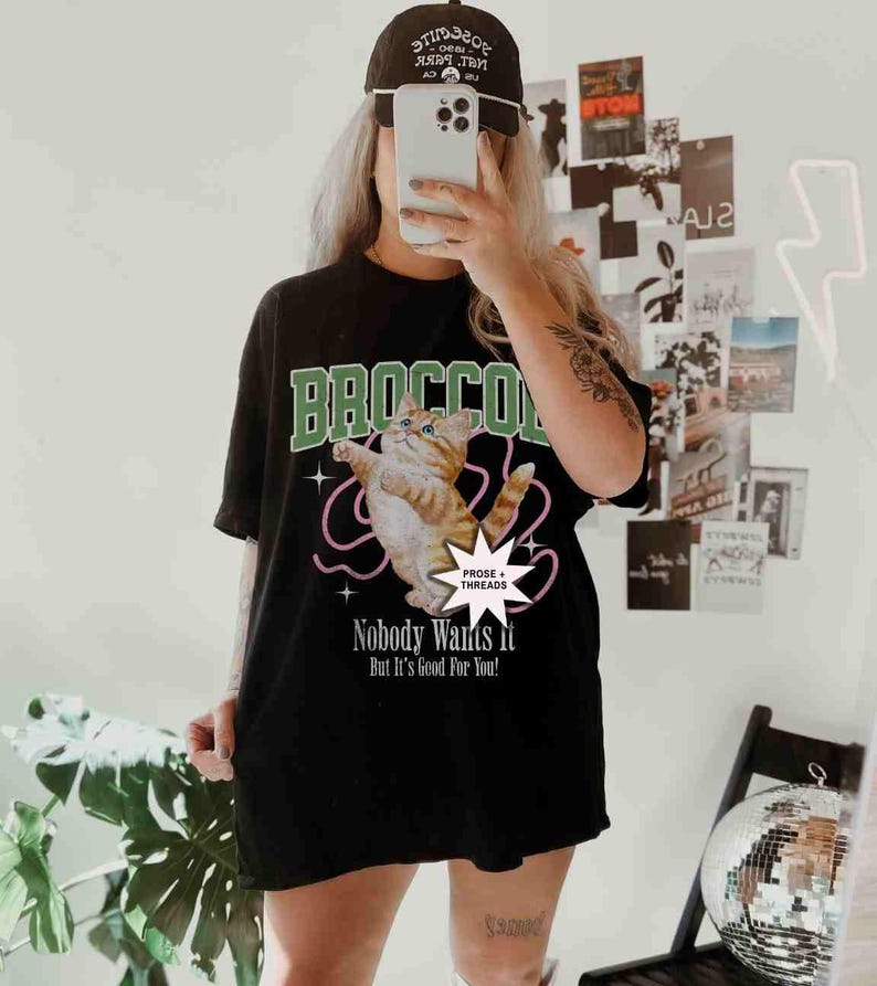 May include: Black oversized t-shirt with a graphic of a cat in a green and white "BROCCO" design. The text "PROSE + THREADS" and "Nobody Wants it But It's Good For You!" is printed on the shirt.