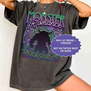 Op de afbeelding: Donkergrijs t-shirt met een grafisch ontwerp met de tekst "MONSTER Romance BOOK CLUB '26". Het ontwerp bevat een silhouet van een monster en decoratieve krullen in paars, groen en blauw. Het shirt heeft ook de tekst "ONLY AT PROSE + THREADS NOT ON TIKTOK SHOP OR SHEIN".