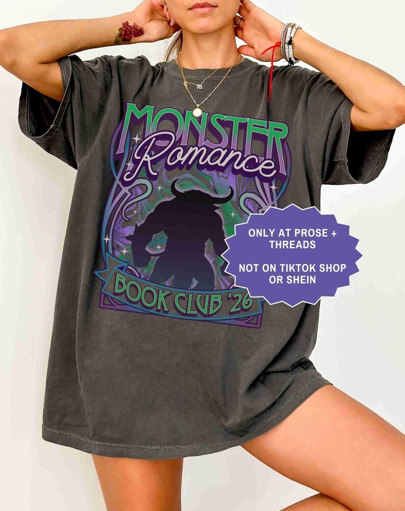 Op de afbeelding: Donkergrijs t-shirt met een grafisch ontwerp met de tekst "MONSTER Romance BOOK CLUB '26". Het ontwerp bevat een silhouet van een monster en decoratieve krullen in paars, groen en blauw. Het shirt heeft ook de tekst "ONLY AT PROSE + THREADS NOT ON TIKTOK SHOP OR SHEIN".