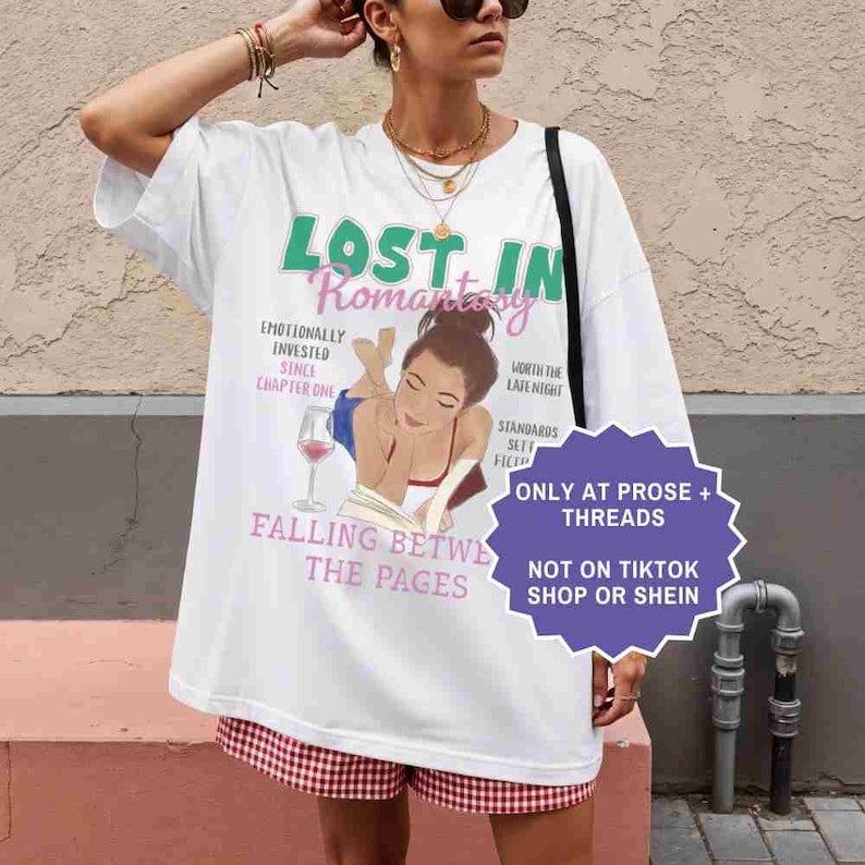 May include: White oversized t-shirt with the text "LOST IN Romantasy" in green and pink. The shirt features an illustration of a woman reading with a glass of wine. Additional text includes "Falling Between the Pages".
